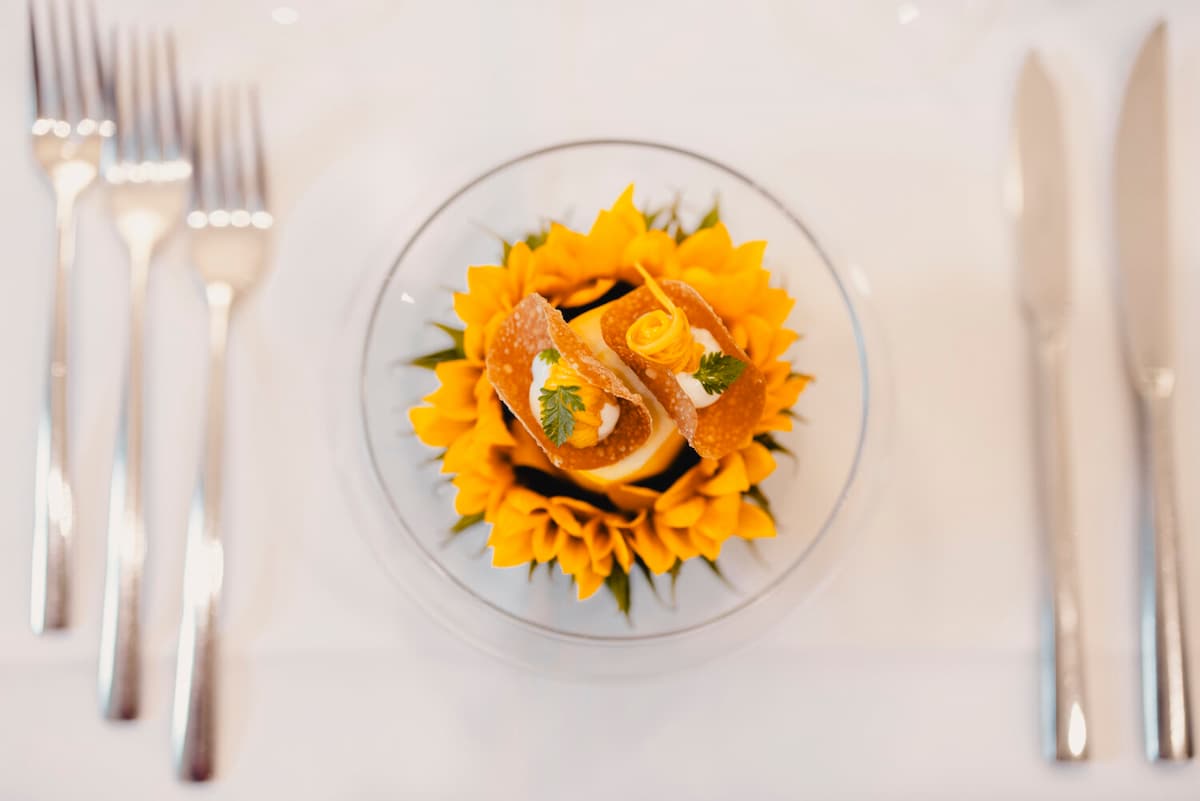 Fine dining dish: golden sunflower-shaped pasta crowned with crispy tuiles, fresh herbs, and a delicate lemon curl on white china.