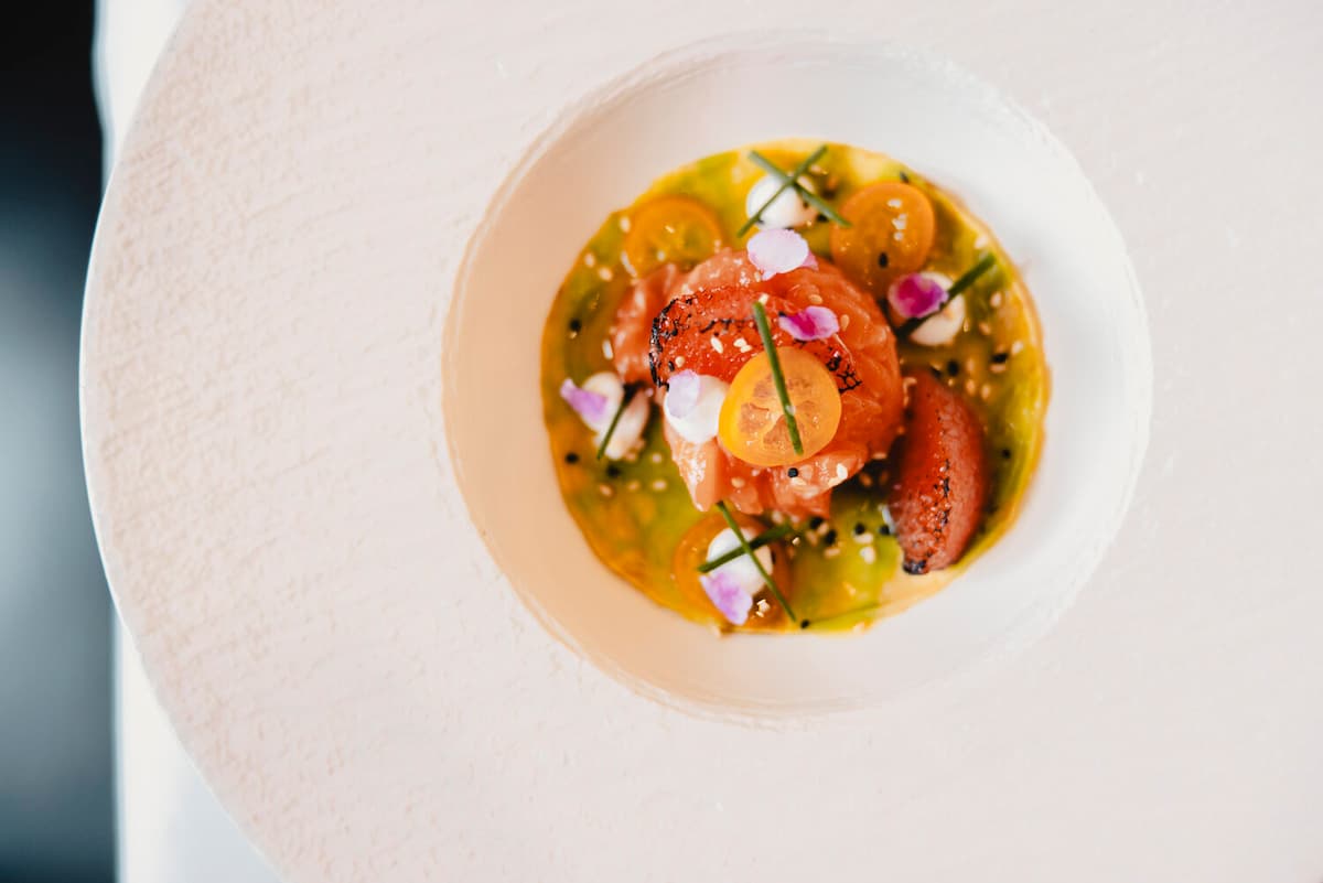 Cured salmon artfully plated in herb-infused oil with cherry tomatoes, edible flowers, and fresh herbs in a white bowl.