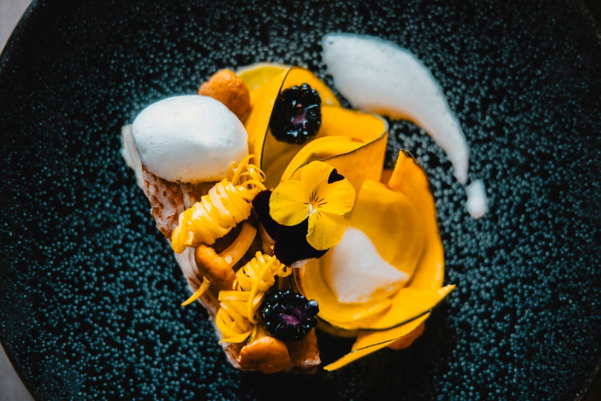 A refined dish arranged on a dark plate — golden petals of pumpkin, a light foam, edible flowers, and berries combine in a vivid contrast of color and texture.
